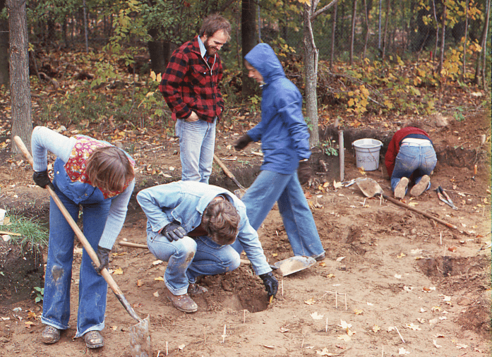 A 1978 field school image showing five archaeologists, three excavating and two standing. One of the standing people appears to be the teacher. Straws are barely visible marking the features uncovered by the students.