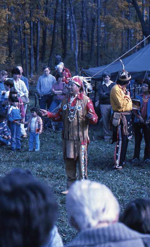 Jim Skye, in full Indigenous regalia stands speaking in the mid-ground of this image of the 1984 Harvest Festival on the Lawson Site. Onlookers and other Indigenous participants surround him.