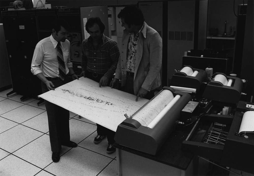 Three men stand over a large print-out of a longhouse feature plan in this black and white image of early technology at the Museum of Ontario Archaeology