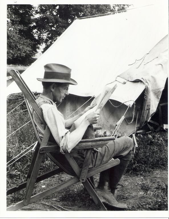 A bespectacled Wintemburg seated in a wooden folding chair in front of a canvas tent, reading the newspaper.