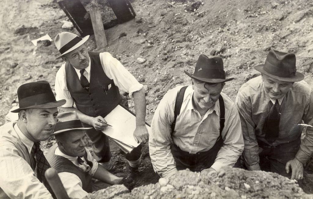 Five men in hats and early 20th century attire consider an archaeological profile outside of the camera frame. One is holding a pencil and paper seemingly to draw the profile.