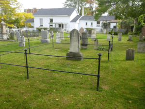 A cemetery plot outlined by a metal chain and fence. The plot contains a headstone and several other grave markers. It is surrounded by other markers. An old church is visible in the background.
