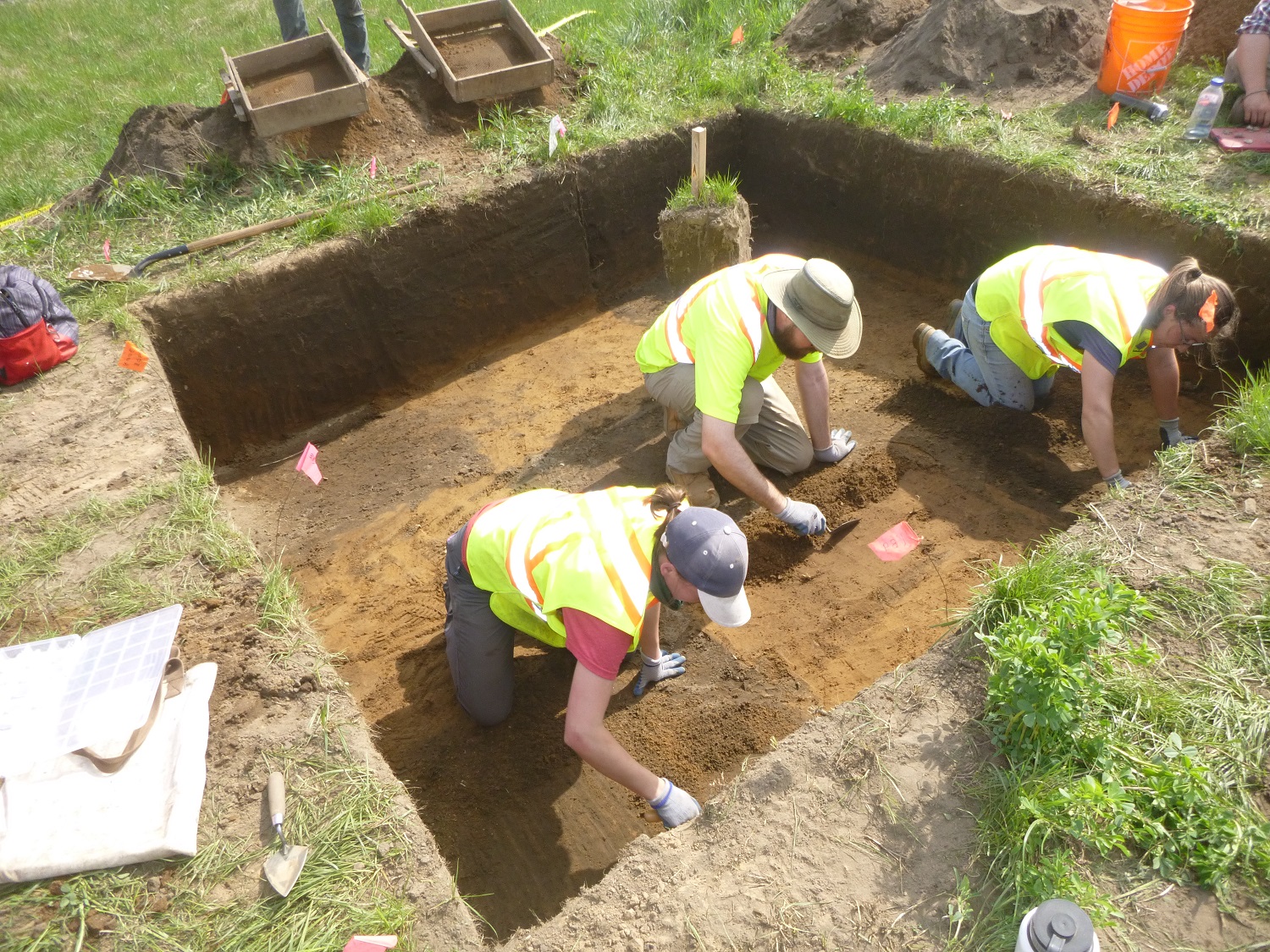Three archaeologists use trowels to investigate the bottom of a Stage 4 excavation.