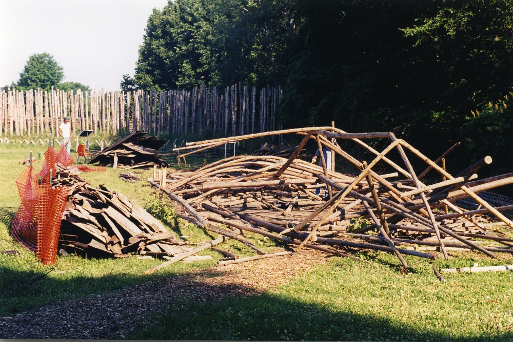 The demolished remains of the larger longhouse on the Lawson Site. A pile of log structural remains is to the right of bark siding debris. Two workers are visible considering the scene