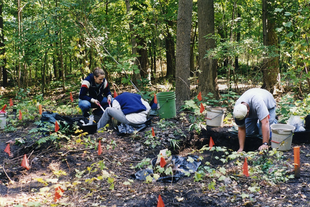 Three field school students kneel next to excavations units. They are surrounded by flags, buckets, and other tools and materials.