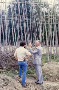 Two men (Finlayson and Wright) stand in front of the Lawson Site's original palisade during construction. Wright's arm is raised as if to show the angle of the palisade as he talks to Finlayson.