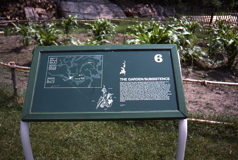 A green interpretive sign titled "The Garden/Subsistence" with a large number 6 and a paragraph illegible text beneath. A map and image of vegetation is also included. The recreated garden at the Lawson Site is visible in the background.