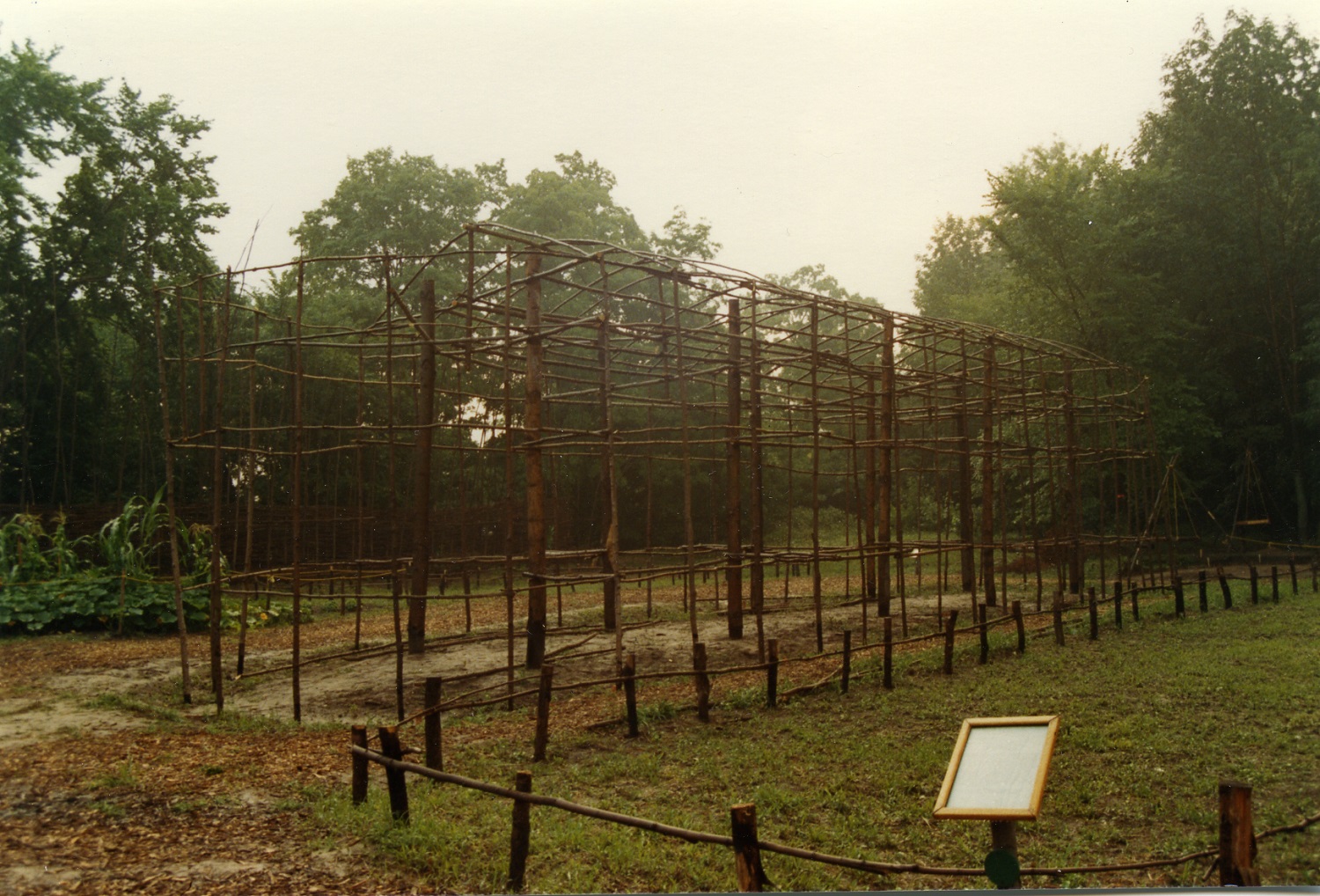 A hazy image of the 1980s frame of the reconstructed longhouse at the Lawson Site. The foreground also shows an outline of another longhouse marked with stakes and saplings in the grass. A plaque or sign is also visible but no legible text can be made out.