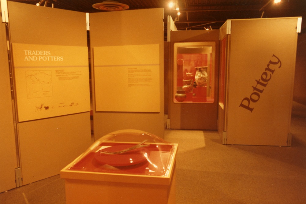 The original Museum of Ontario Archaeology gallery as it appeared in the 1980s. A panel labelled "Traders and Potters" is mounted beside a display case featuring pottery. The word "Pottery" appears next to the case. An artifact is displayed on a pedestal in the foreground