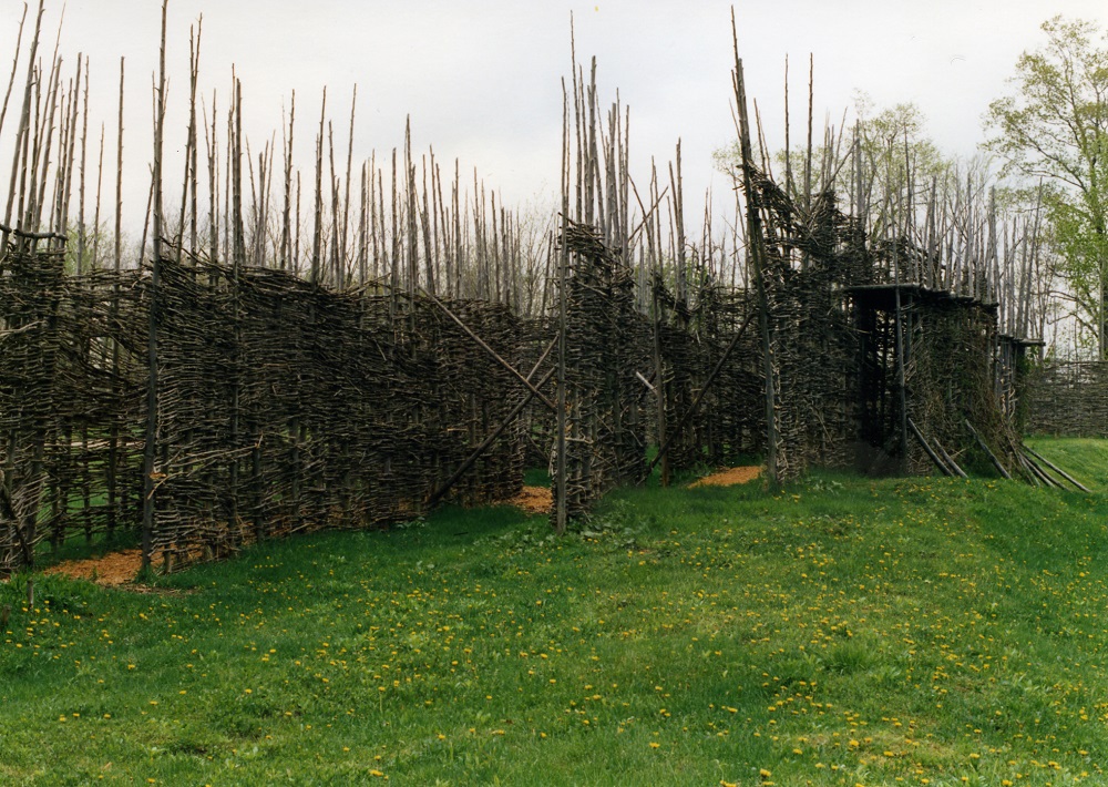 The original palisade at the Lawson Site in the 1990s. A latticework of saplings attached to vertical poles form a maze with raised interior platforms. Grass surrounds the walls.