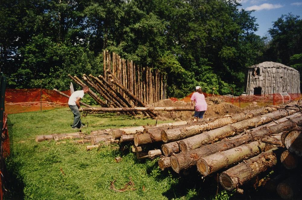 Two workers carry a log pole to its place in the 2000 reconstruction of the Lawson Site palisade. The image shows the various stages of pole installation. Erected (background), placed (middle) and a log pile in the foreground. The bark-covered longhouse is also visible in the background.