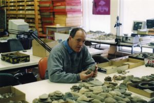 A man (Bob Pearce) looks up at the camera from considering a stone artifacts which he holds in his hands. Around him are tables covered in artifacts and boxes of artifacts. Shelves of wood artifact trays are visible in the background