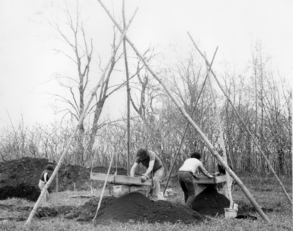 Three workers hunch over screens sifting dirt in this 1980 image of Lawson Site excavation. A fourth is visible in the background with a shovel and piles of dirt.