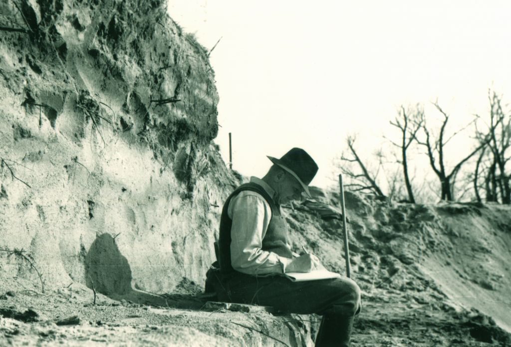 A man (Wilfrid Jury) sits on an excavated earth bench with a deep strata behind him. He is marking a notebook.
