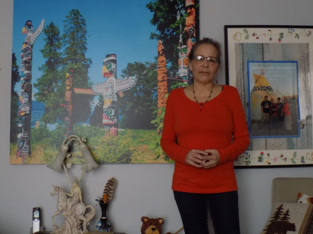 An Indigenous person(Wanda Maness) stands in front of a wall featuring a picture of totem poles and a framed poster/magazine cover.