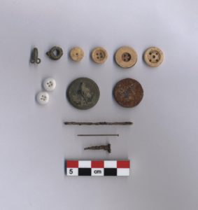 A assorted set of historical artifacts including buttons, coins, nails, and grommets. A scale bar is shown beneath.