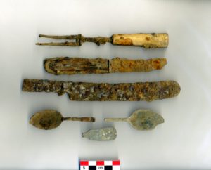 A set of historical utensil artifacts including two knives, one fork, and three spoon fragments. A scale bar is shown underneath