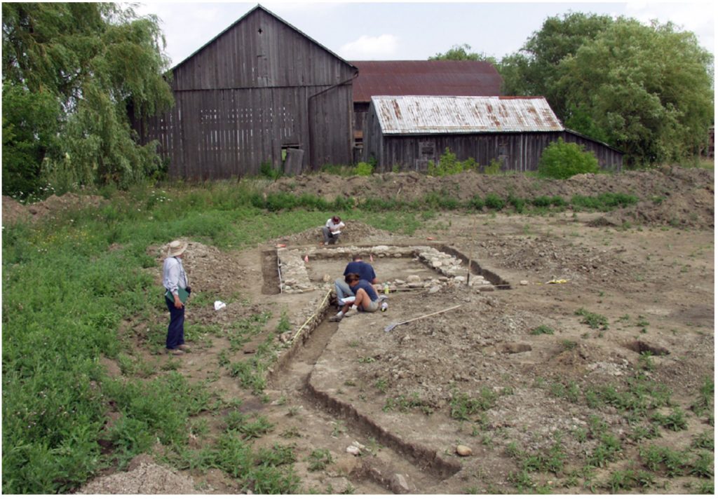 In the foreground, four archaeologists observe and work on an exposed stone foundation in a previously stripped section of a property. Several old barns are visible in the background.