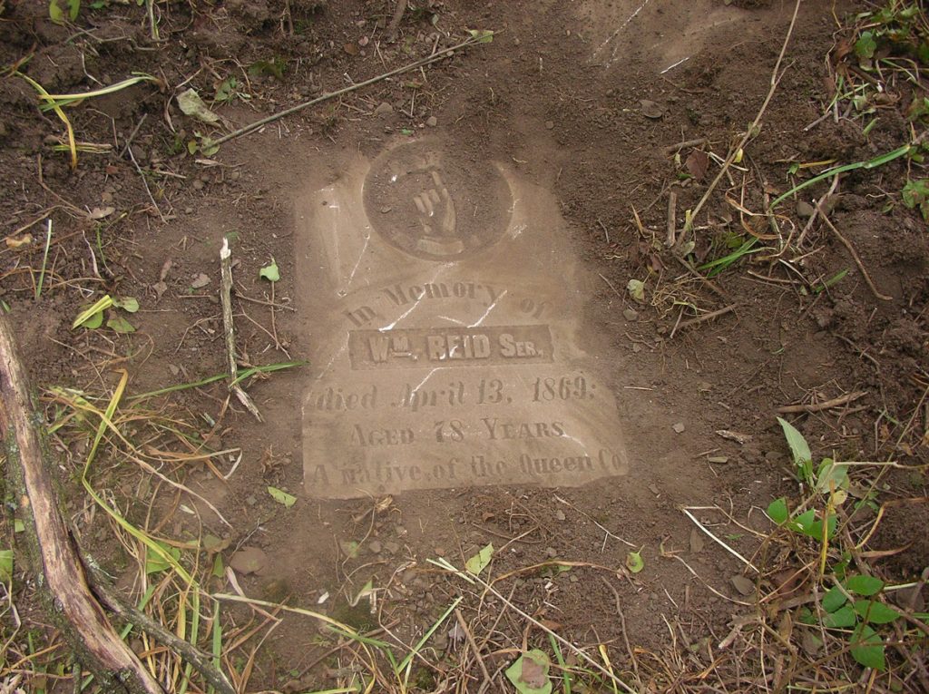 A gravestone is shown lying flat and uncovered in the dirt. The words read 