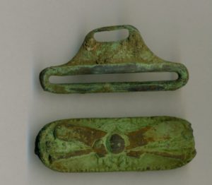 Hardware from a set of cricket (sport) suspenders. The metal is green from age and shows a crossed pair of cricket bats.