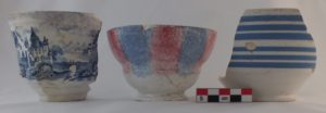 Three artifacts, two cups and a bowl shown in relatively good condition. All are ceramic. The left cup shows a bridge scene in a blue willow motif. The center cup has an alternately red and blue sponged motif extending down from it's lip. The right bowl fragment has six blue bands horizontally.