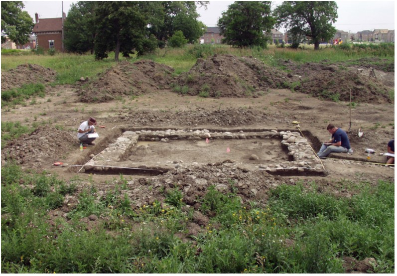 Three archaeologists sit or kneel and record a stone foundation uncovered in an open field. A new subdividision is visible in the distance.