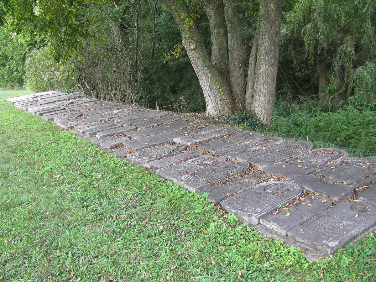 Two rows of displaced headstones are arranged at the edge of a grassy area and a forest.