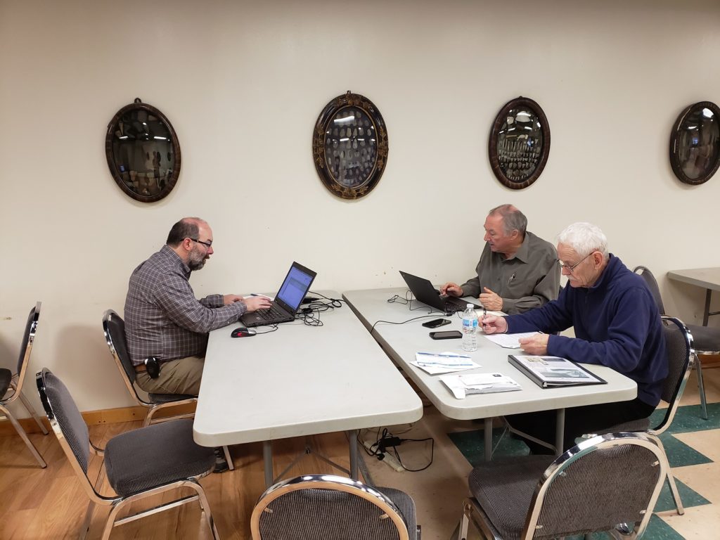 Three volunteers (Steve Naftel, Chris Ellis, Darryl Dann) site around a table working on laptops and reading from papers. Mounted artifacts are on display behind them.