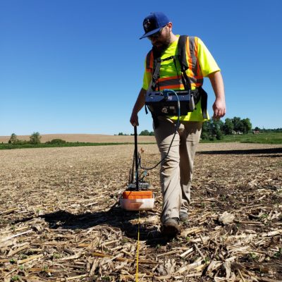 Tom Porawski pulls the Ground Penetrating Radar unit over an old corn field towards the camera looking up at him against the blue sky.