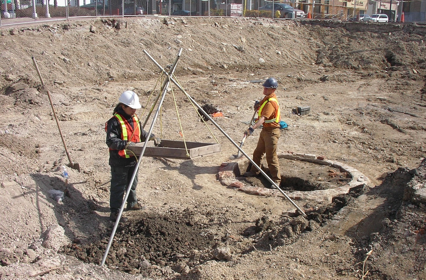 Two archaeologists stand in the bottom of a large excavation in an urban setting. One works the screen while the other digs out an old well or cistern.