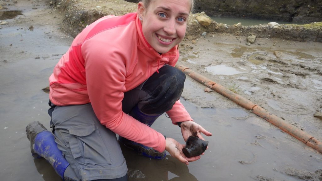 Alex Kisielewski kneels in a wet excavation holding an artifact in her hands and looks at the camera smiling