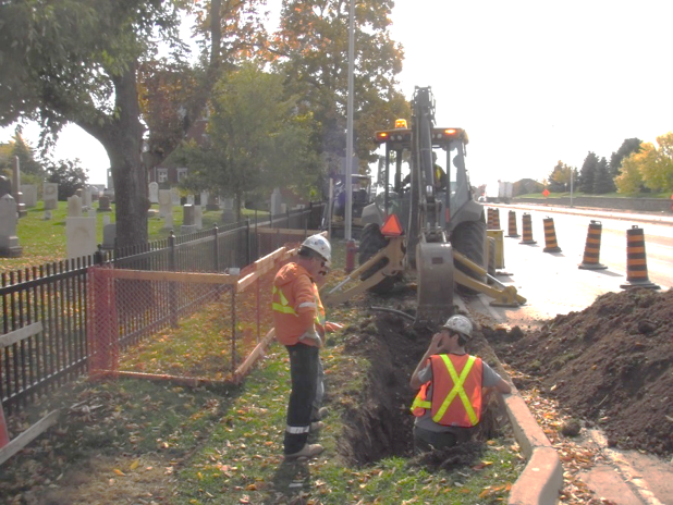 A worker examines a trench in front of a bachoe while another looks on.